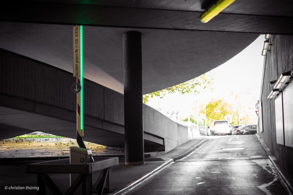 Betonrampe mit Schranke und Leuchtsignal, Autos im Hintergrund, Tageslicht.