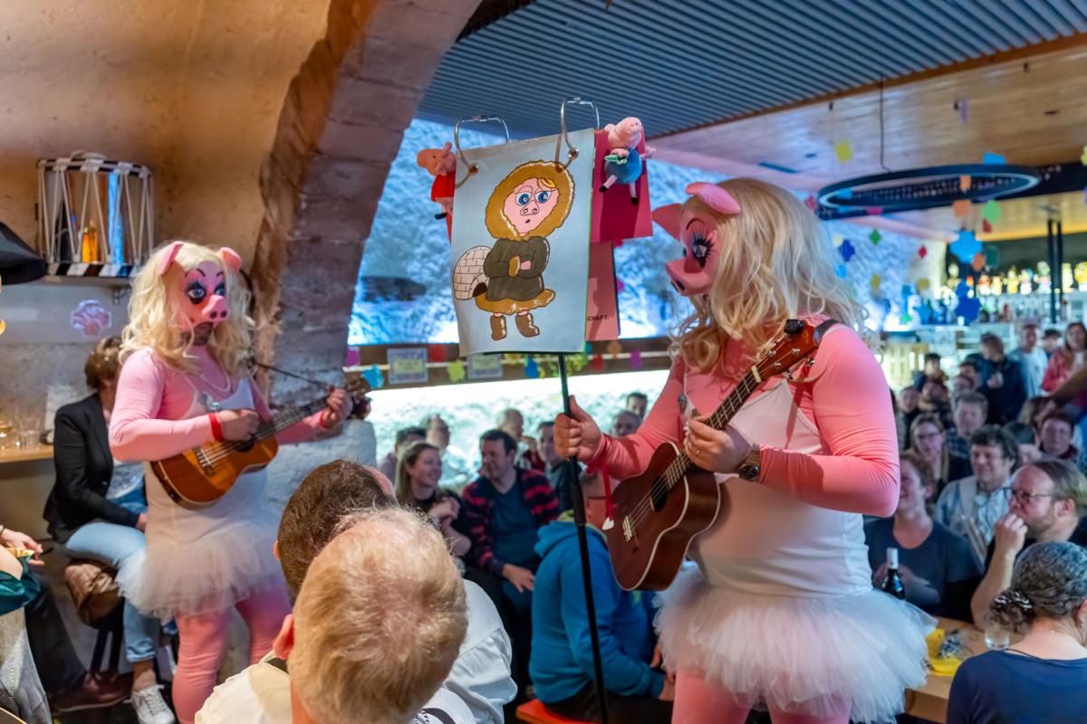 Zwei Personen mit Schweine-Larven und rosa Kostümen spielen Ukulele in einem vollen Lokal, Publikum lacht.