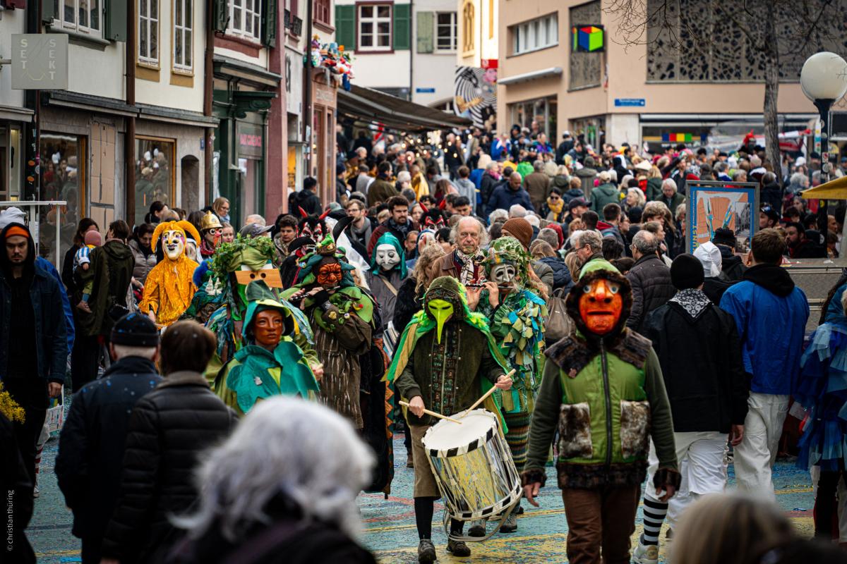 Fasnachtsgruppe mit grünen Larven und Trommler zieht durch eine belebte Gasse, umgeben von Zuschauern.
