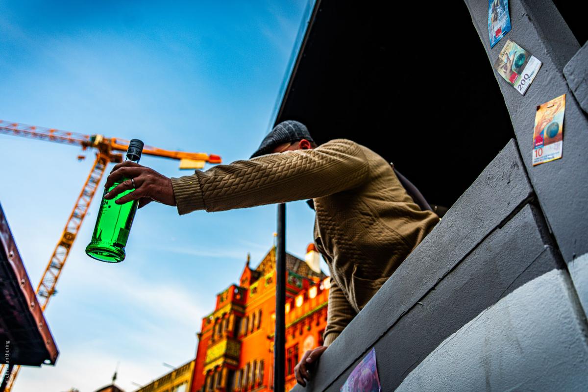 Person reicht grüne Flasche aus einem Wagenfenster, Baukran und Altstadt im Hintergrund.