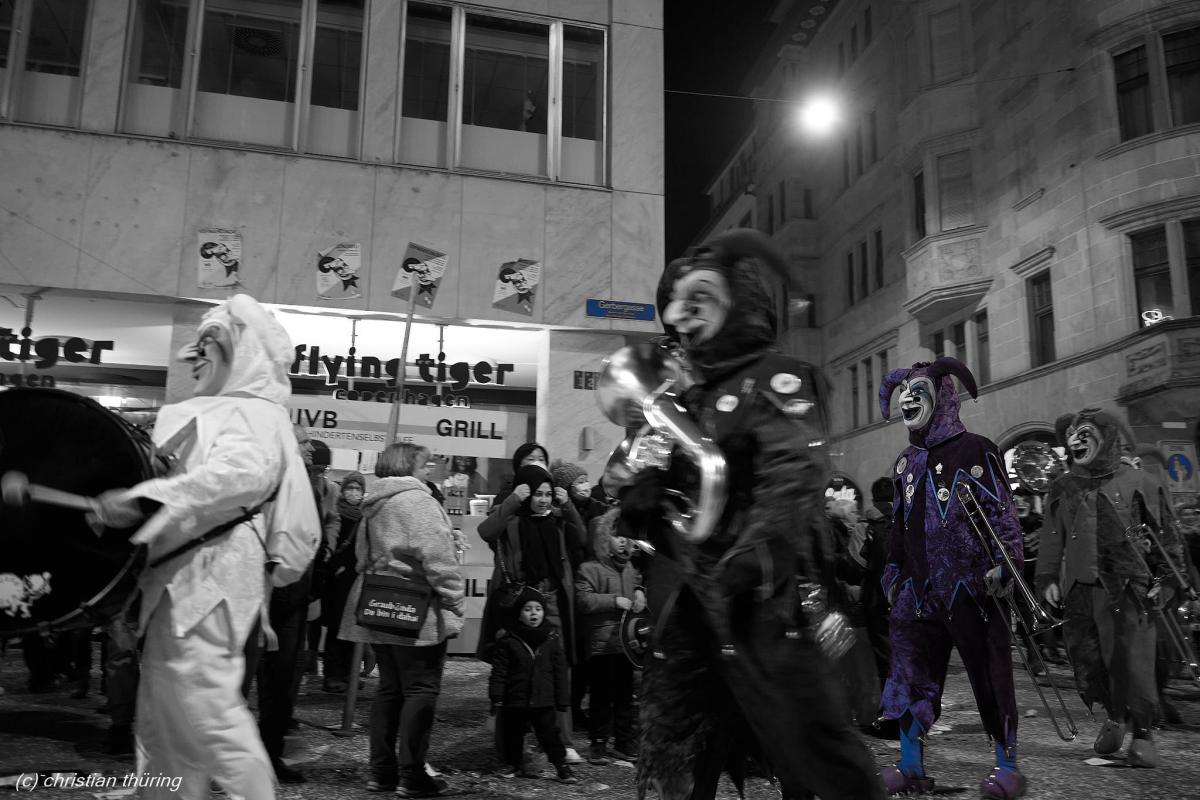 Mehrere kostümierte Fasnächtler mit Larven und Instrumenten ziehen nachts durch eine Basler Straße.
