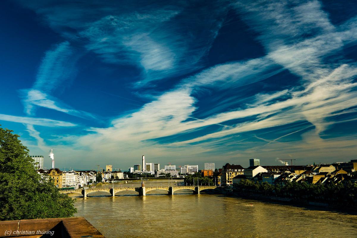Blick auf eine Brücke über den Rhein mit Stadtgebäuden und auffälligen Wolken am Himmel.