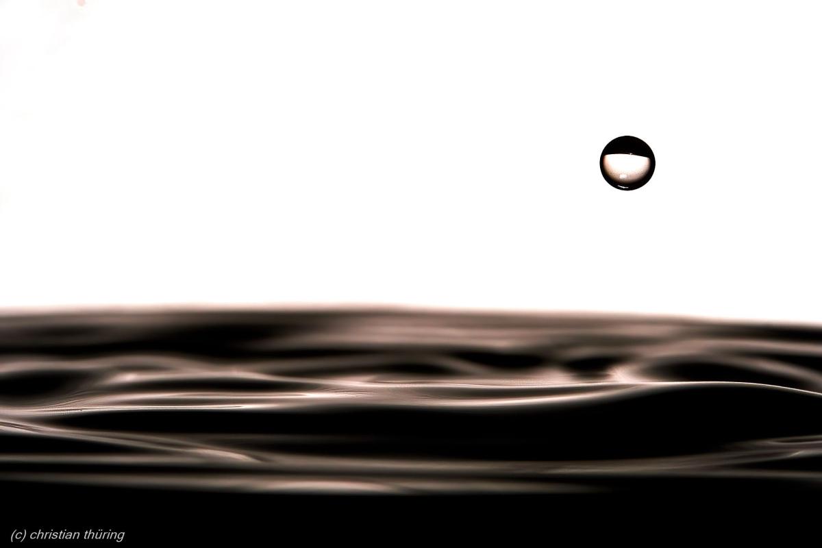 Ein einzelner Wassertropfen schwebt über einer leicht bewegten Wasseroberfläche vor hellem Hintergrund.