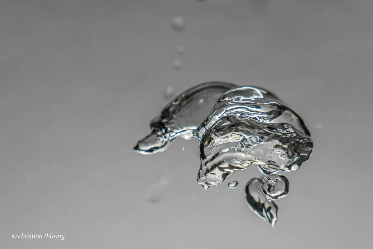 Eine einzelne Wasserblase schwebt in transparenter Flüssigkeit vor grauem Hintergrund.