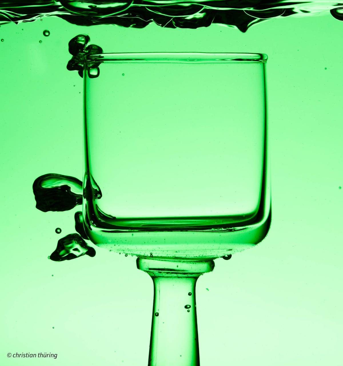 Ein quadratisches Glas steht kopfüber in grünem Wasser, Luftblasen steigen auf.