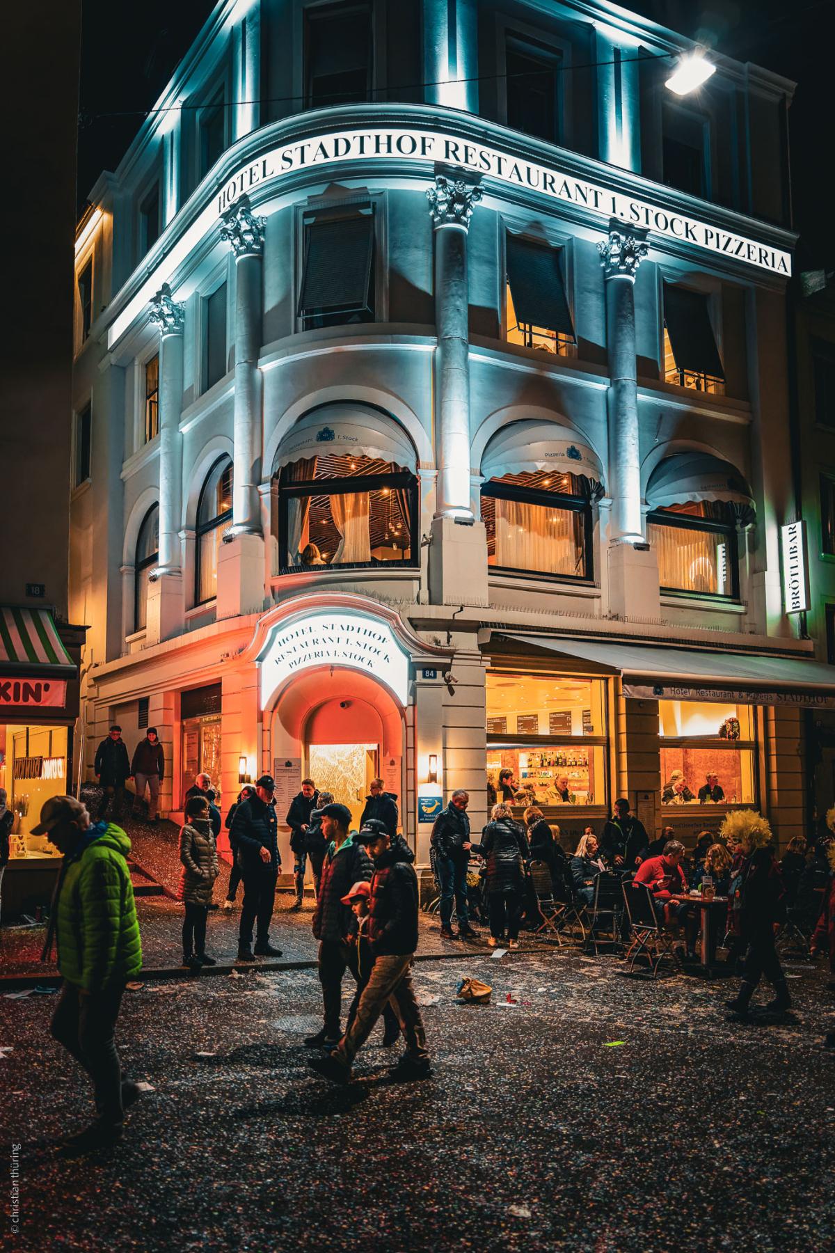 Menschen stehen und sitzen abends vor dem hell erleuchteten Hotel Stadthof, Konfetti auf der Straße.