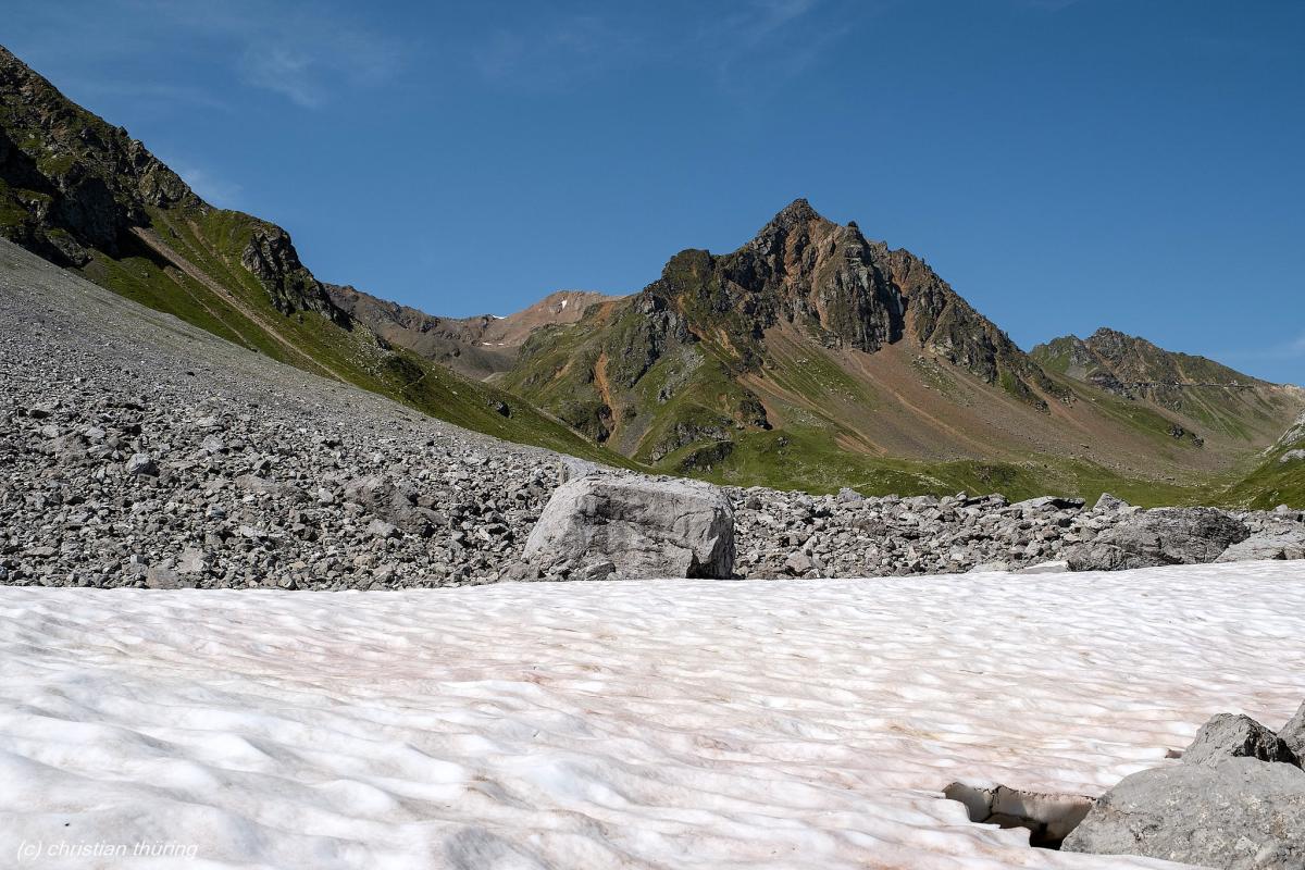 Felsige Berggipfel mit grün-braunen Hängen, Geröllfeld und Schneefeld im Vordergrund unter blauem Himmel.