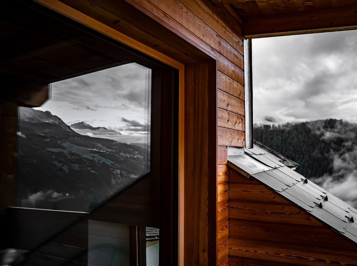 Holzverkleidete Hausfassade mit Fenster, das Berge und Wolken reflektiert. Nebel liegt über bewaldeten Hügeln.
