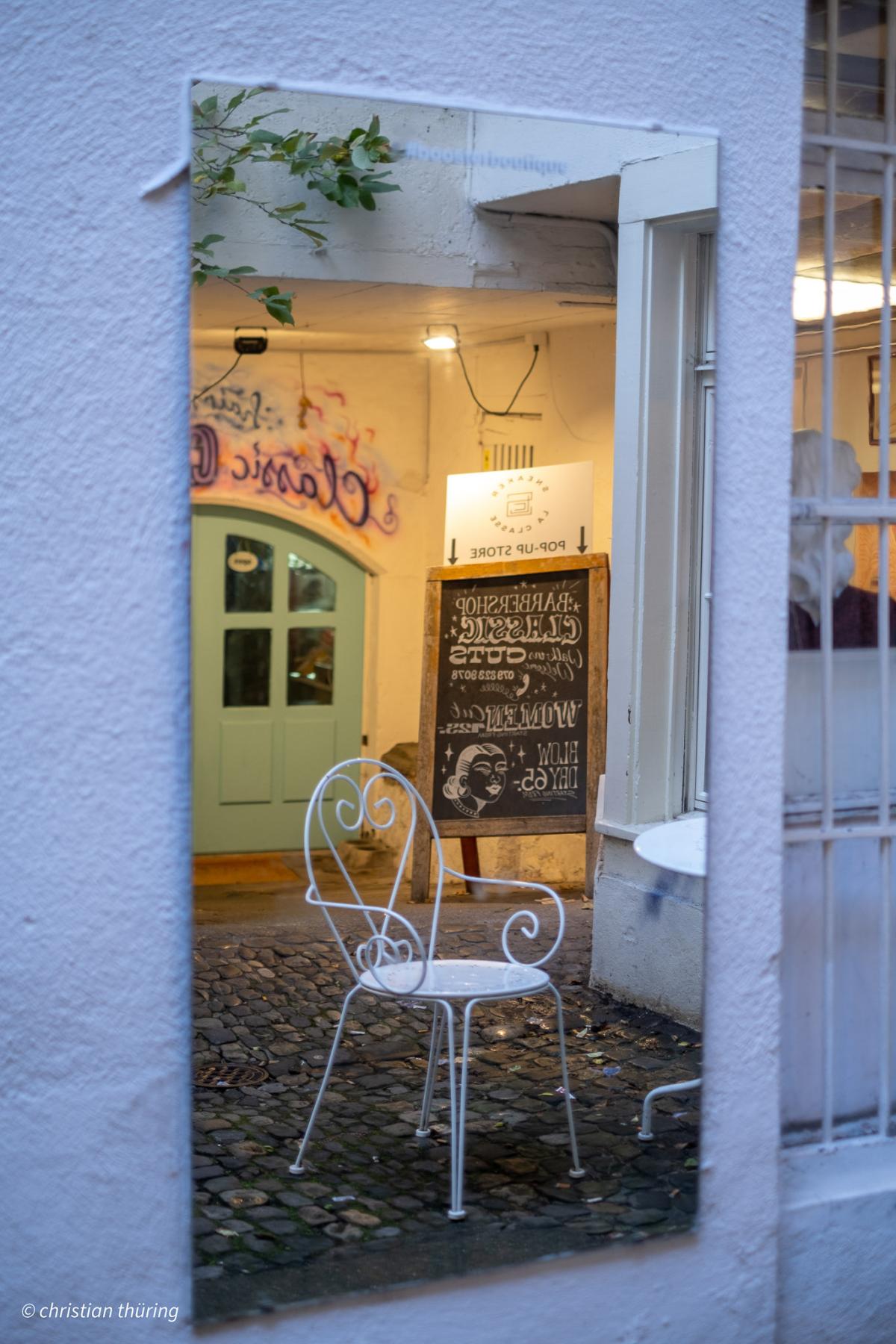 Ein weißer Metallstuhl steht auf Kopfsteinpflaster und spiegelt sich in einem Wandspiegel, im Hintergrund ein Pop-up-Store.