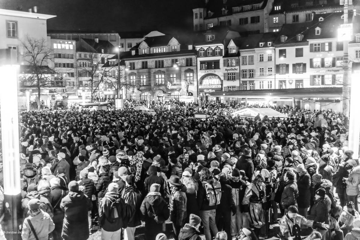 Dichte Menschenmenge auf einem beleuchteten Platz vor historischen Häusern bei Nacht.