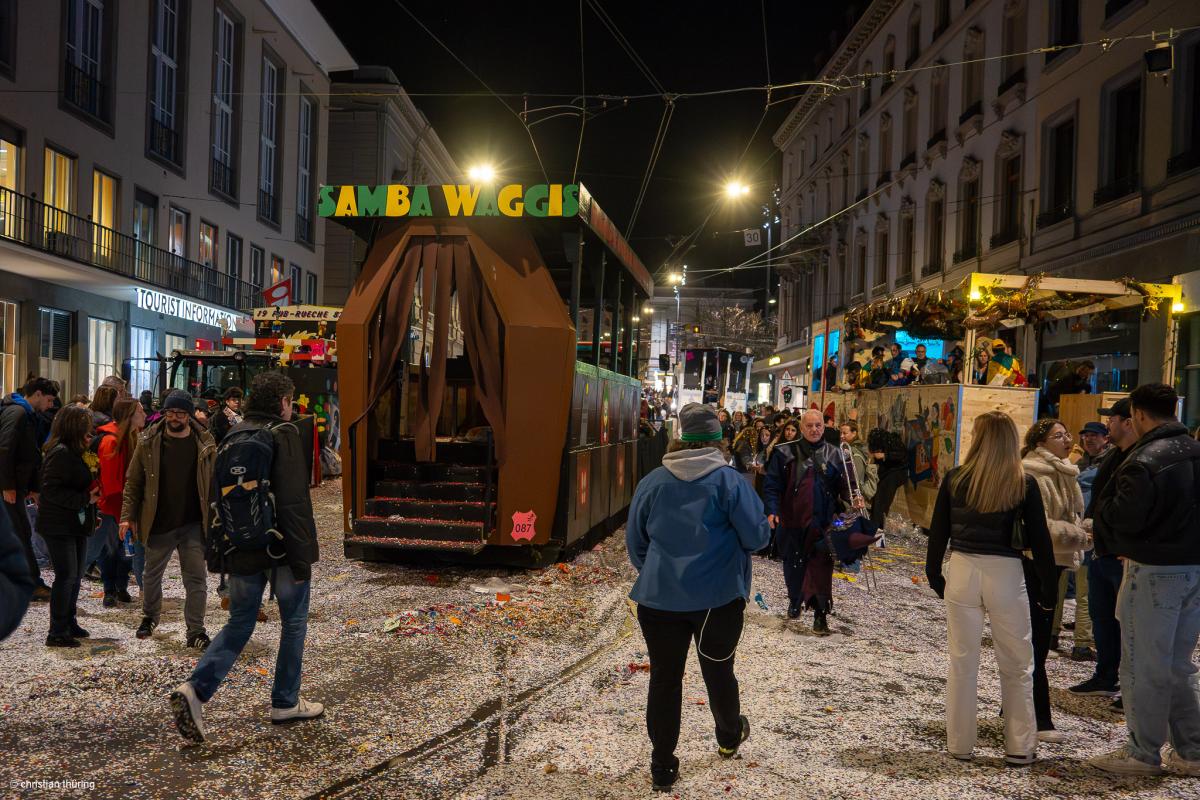 Ein geschmückter Fasnachtswagen mit der Aufschrift Samba Waggis steht nachts in einer Straße, umgeben von Menschen und Schyssdräggziigli.