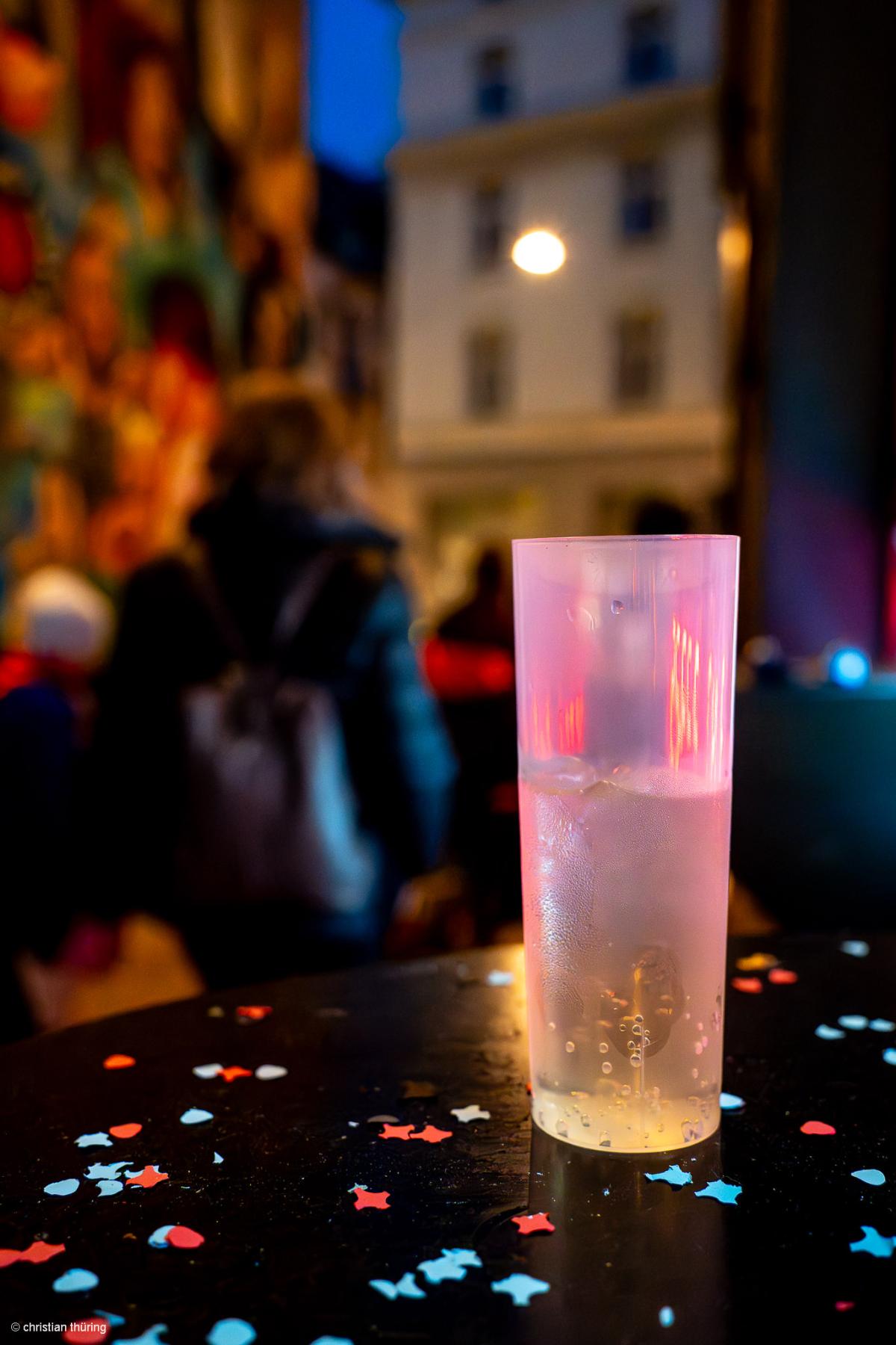 Ein halbvoller Plastikbecher mit Eiswürfeln steht auf einem runden Tisch mit buntem Konfetti, im Hintergrund unscharfe Menschen.
