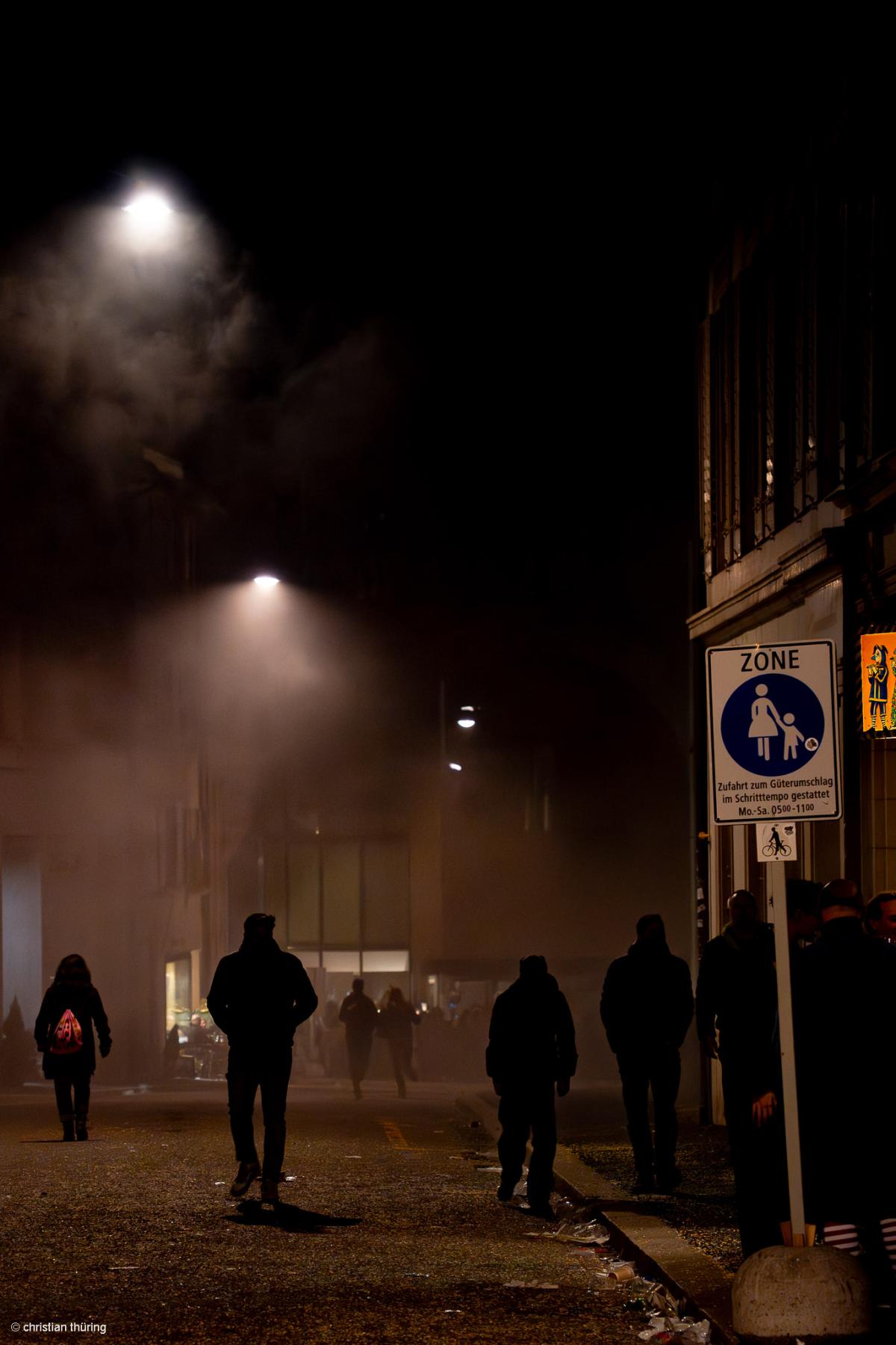 Mehrere Personen gehen nachts durch eine von Nebel und Straßenlaternen beleuchtete Gasse.