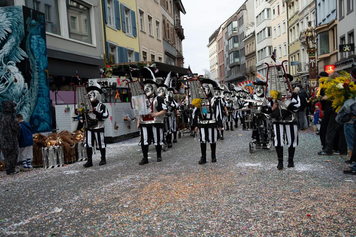Eine Clique in schwarz-weiss gestreiften Kostümen mit Larven und Glockenspielen marschiert durch eine mit Schyssdräggziigli bedeckte Straße.