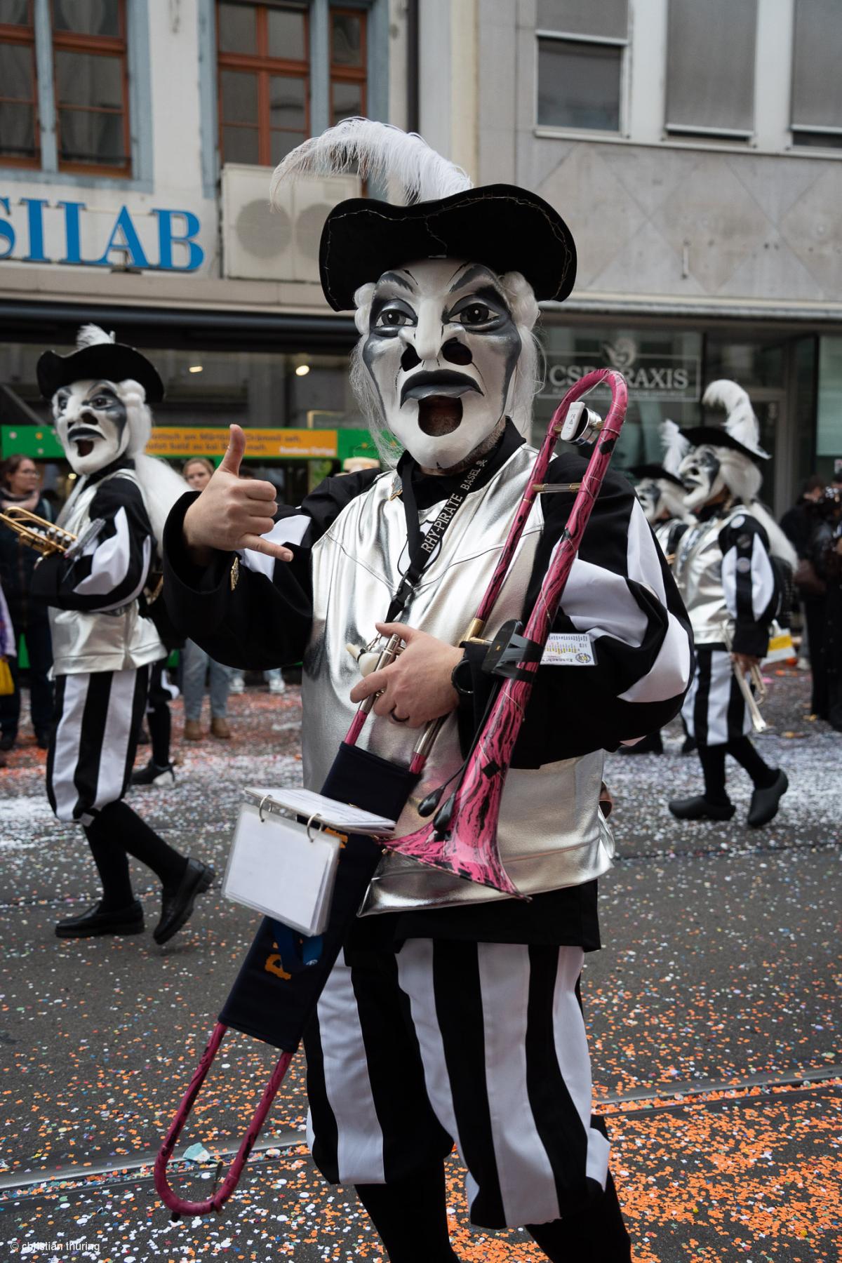 Ein Mitglied der rhy-pirate Clique mit schwarz-weisser Larve und Posaune, umgeben von Schyssdräggziigli auf der Straße.