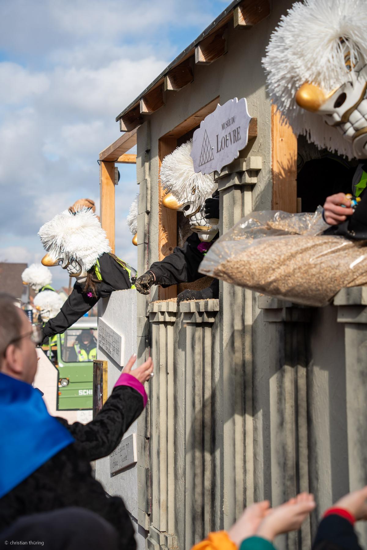 Mehrere Waggis mit weissen Larven verteilen Schyssdräggziigli von einem Fasnachtswagen an Zuschauer.