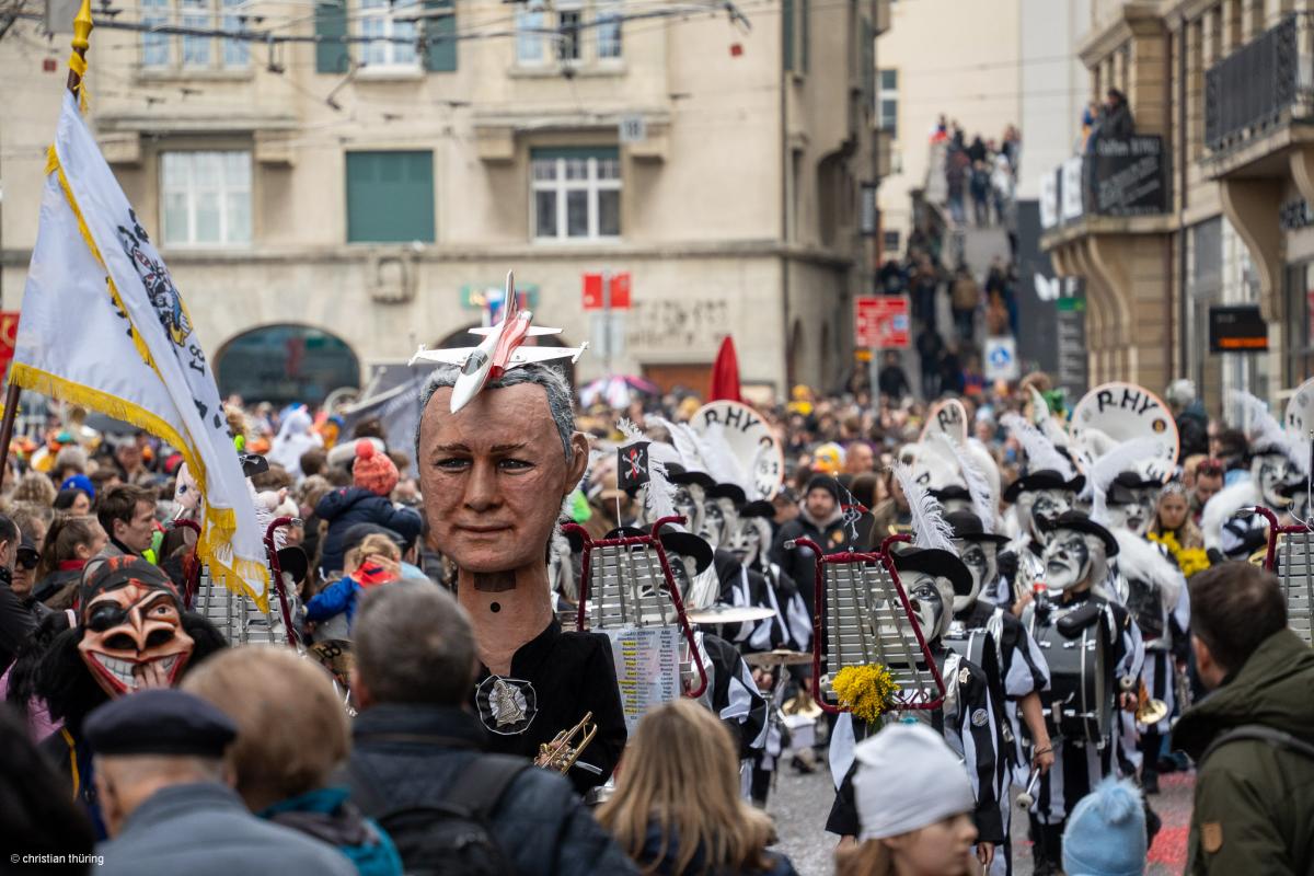 Große Basler Fasnachtsgruppe mit weissen Larven, Instrumenten und Fahne in einer vollen Innenstadt.