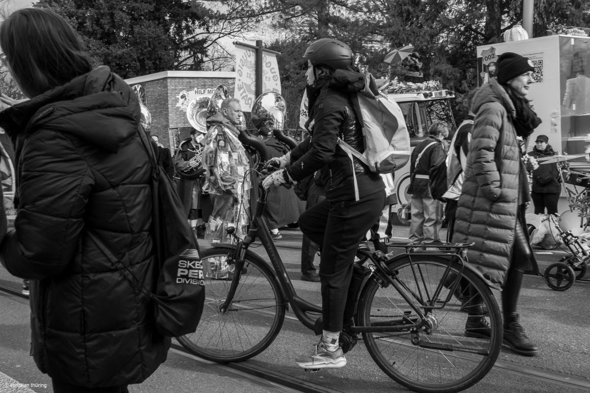 Schwarzweissfoto mit Guggemuusig, Menschen in Winterjacken und einer Person auf dem Velo während des Basler Cortege.